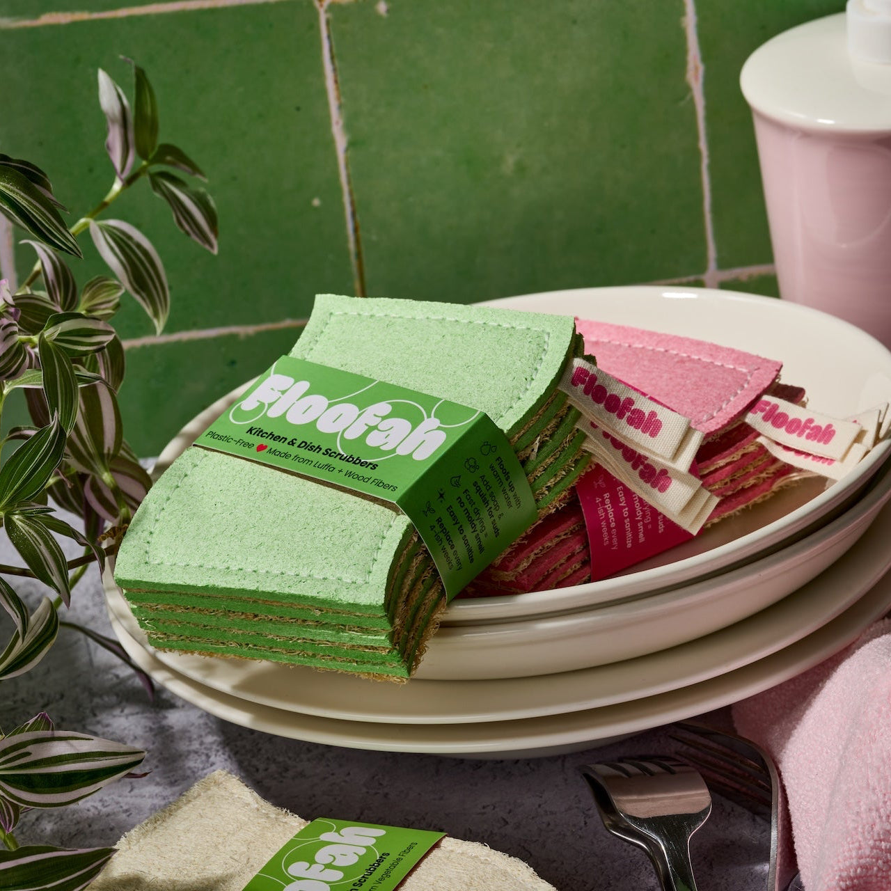 A stack of green and pink kitchen sponges with visible 'Floofah' branding, arranged on a white plate in a styled kitchen-like setting with green tiled walls, a pink pitcher, and a pink towel.
