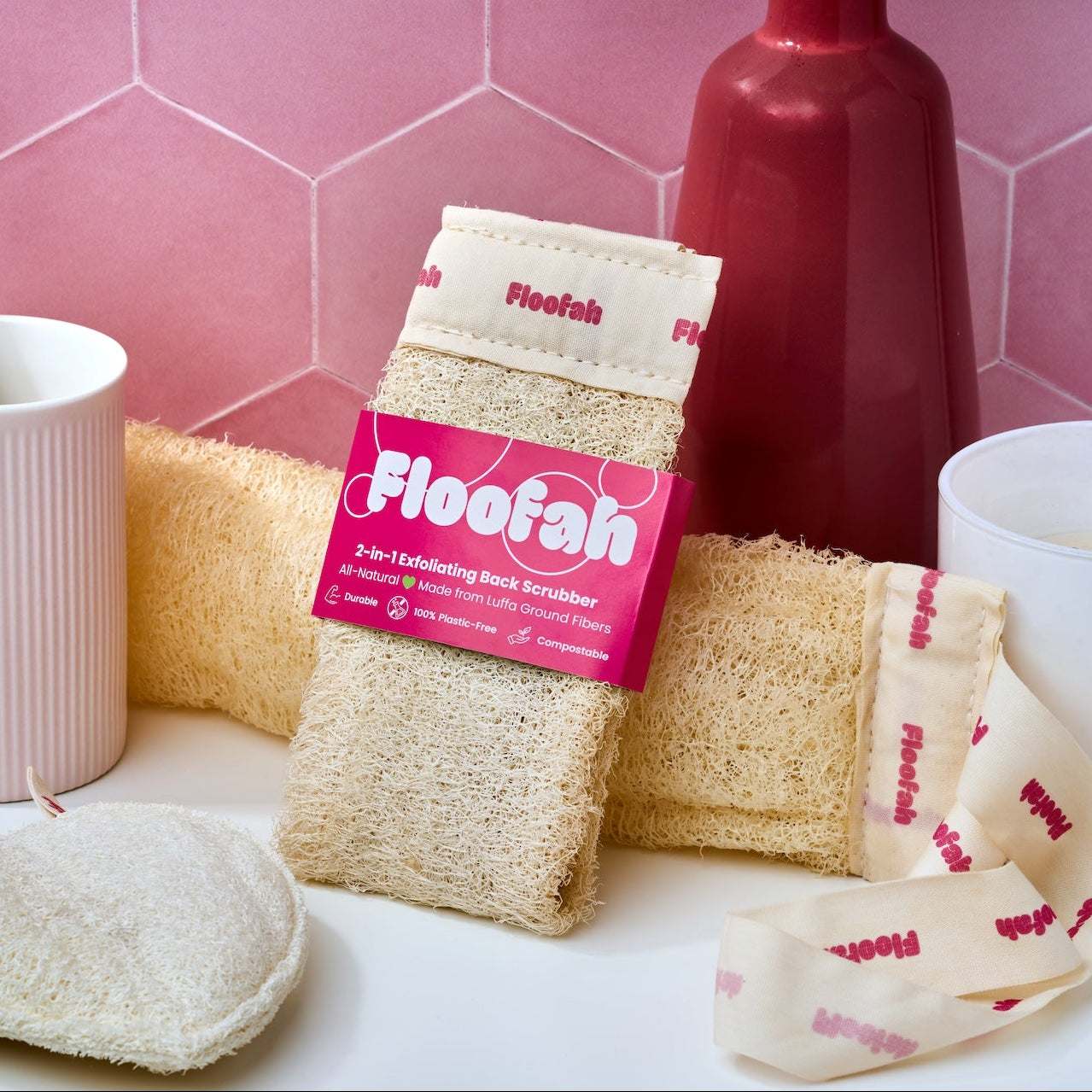 Floofah plant shower sponge and back scrubber shower tool displayed on a white surface against a pink hexagonal tile background, including packaged sponge washcloths, loose beige sponges, a pink mug with wooden toothbrushes, and a white bowl.